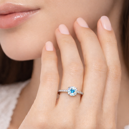 Close-up of a hand wearing a ring with a blue gemstone, against a neutral background.