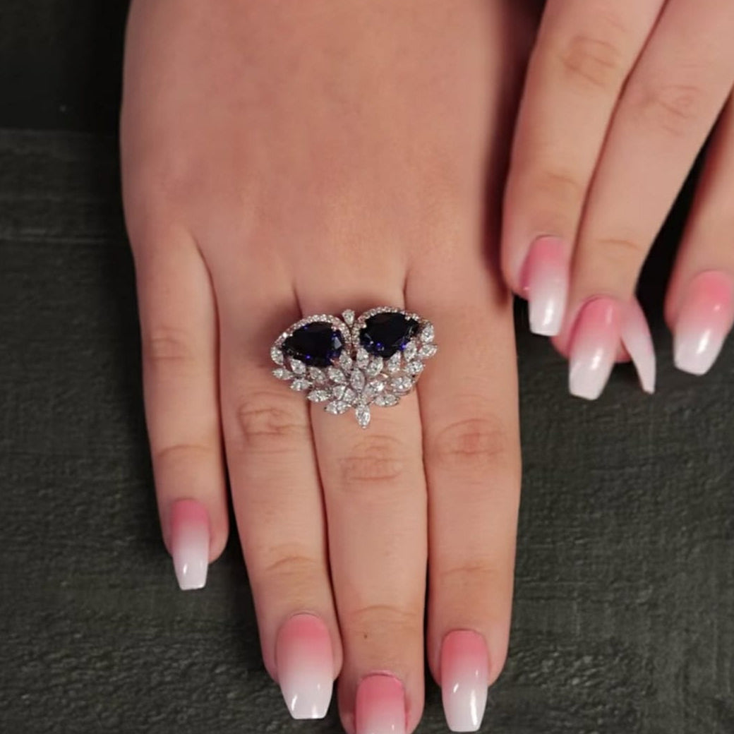Heart-shaped ring with a central gemstone on a hand with pink nail polish.