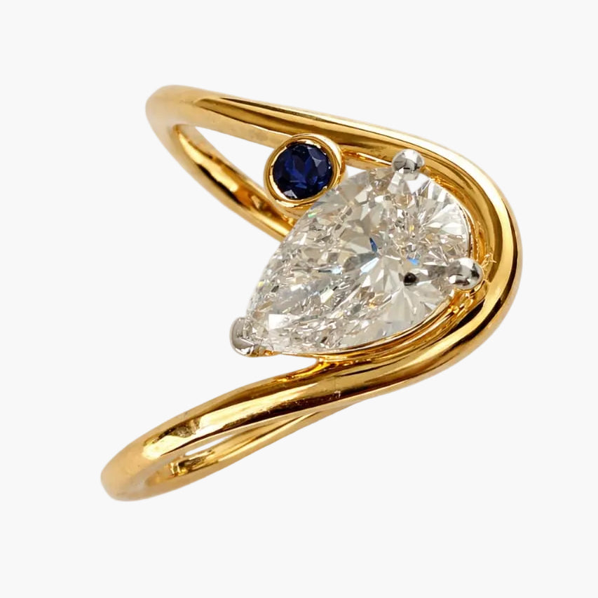 Gold ring with a pear-shaped diamond and a small blue gemstone on a white background