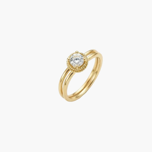 A gold ring with a round-cut clear gemstone in a bezel setting, accented by a halo of small diamonds and a textured outer border, shown on a plain white background.