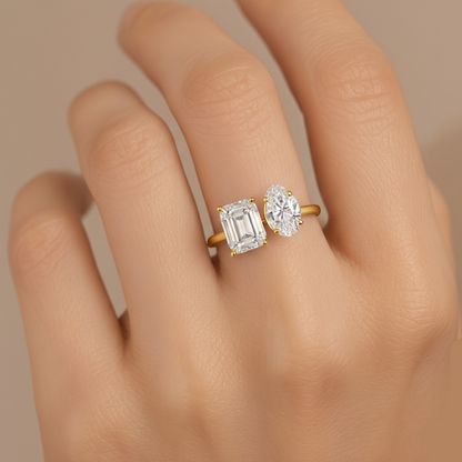 Gold ring with emerald-cut and round diamond on a hand against a neutral background