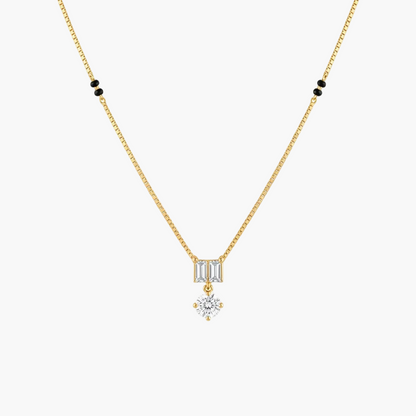 A delicate gold chain necklace featuring a small rectangular baguette-cut clear stone pendant, with a dangling round clear gemstone below, accented by small black beads on the chain against a white background.