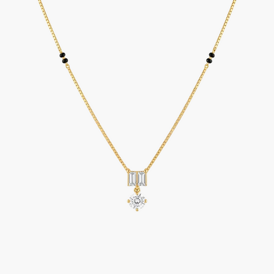 A delicate gold chain necklace featuring a small rectangular baguette-cut clear stone pendant, with a dangling round clear gemstone below, accented by small black beads on the chain against a white background.