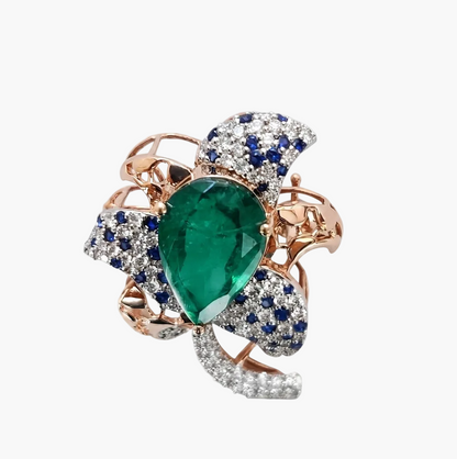 A rose gold ring featuring a large pear-cut green emerald center stone, surrounded by pavé-set blue sapphires and clear diamonds, on a clean white background.
