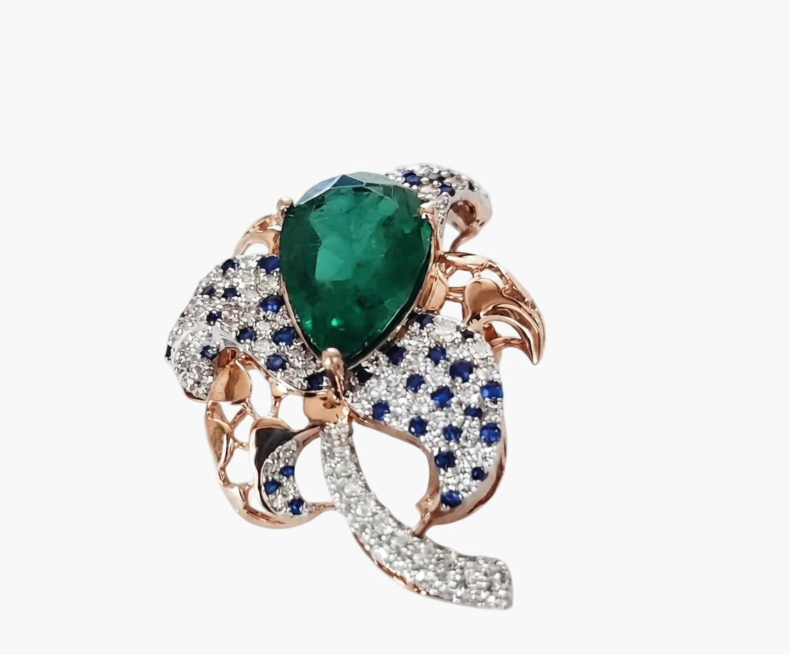 A rose gold ring featuring a large pear-cut green emerald at the center, surrounded by pavé-set blue sapphires and clear diamonds, with curved rose gold and diamond accents on a white background.