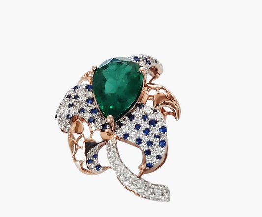 A rose gold ring featuring a large pear-cut green emerald at the center, surrounded by pavé-set blue sapphires and clear diamonds, with curved rose gold and diamond accents on a white background.