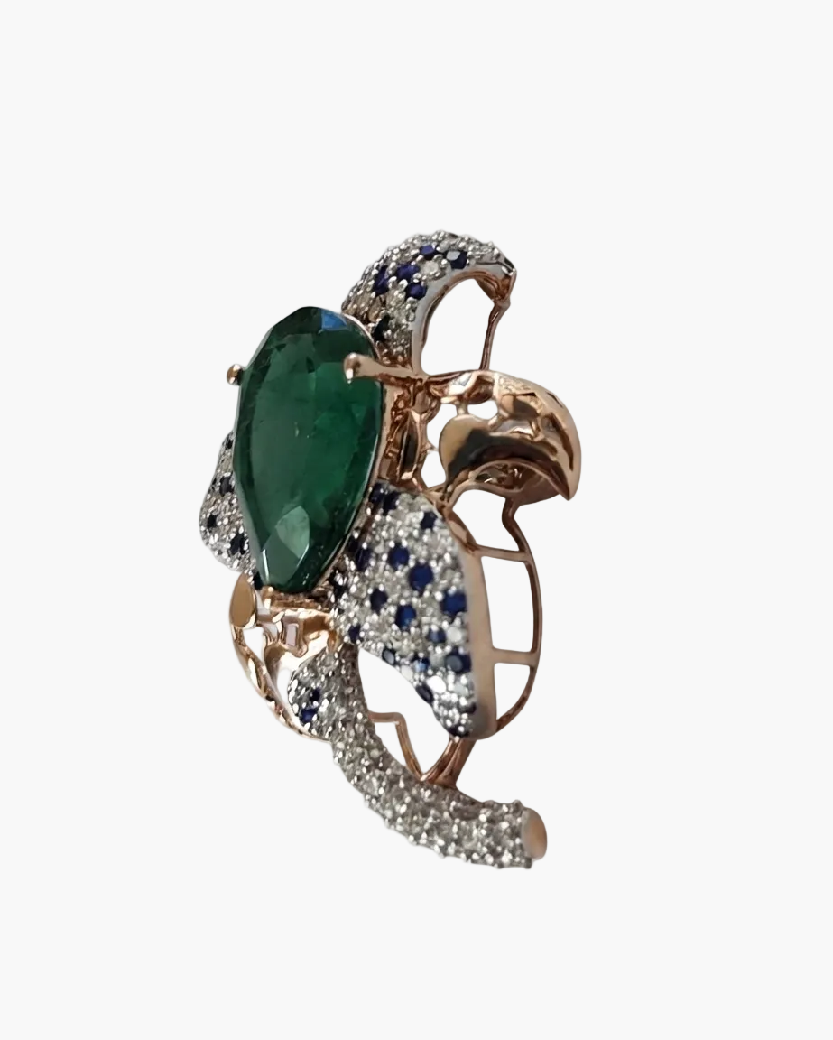A rose gold butterfly ring featuring a large green emerald-cut center stone, surrounded by pavé diamonds and accented with round blue sapphires, set against a plain white background.
