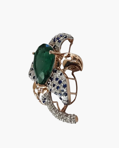 A rose gold butterfly ring featuring a large green emerald-cut center stone, surrounded by pavé diamonds and accented with round blue sapphires, set against a plain white background.