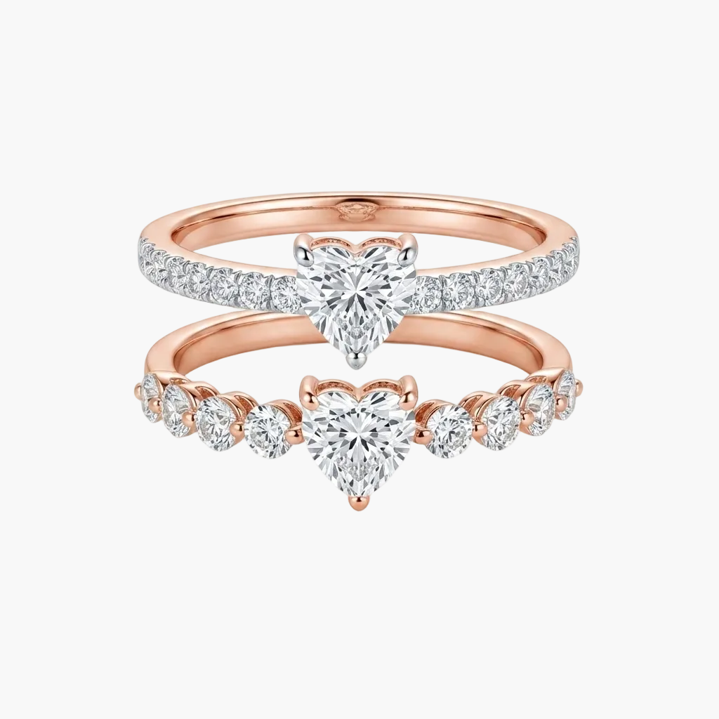 A pair of rose gold wedding rings stacked together on a white background. The top ring features a pavé-set heart-shaped diamond, and the bottom ring features a row of small round diamonds leading to a central heart-shaped diamond.
