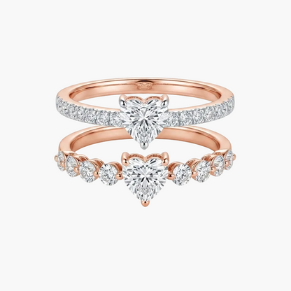 A pair of rose gold wedding rings stacked together on a white background. The top ring features a pavé-set heart-shaped diamond, and the bottom ring features a row of small round diamonds leading to a central heart-shaped diamond.