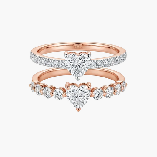 A pair of rose gold wedding rings stacked together on a white background. The top ring features a pavé-set heart-shaped diamond, and the bottom ring features a row of small round diamonds leading to a central heart-shaped diamond.