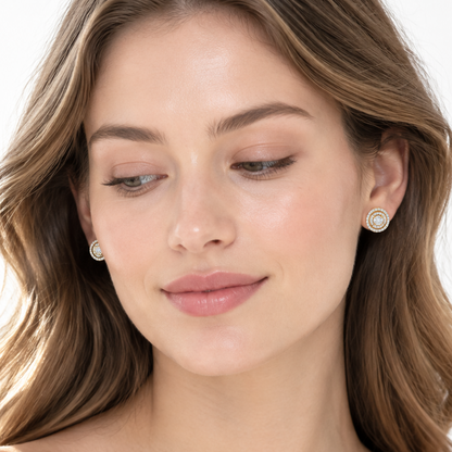 Beautiful elegant female model with light porcelain skin and natural dewy no-makeup look with soft pink lips and soft natural waves loosely tucked behind both ears wearing very small yellow gold double halo round diamond stud earrings each featuring a small round brilliant-cut center diamond in four tiny yellow gold claw prong settings surrounded by a first inner circular pavé diamond halo then a thin plain yellow gold rim and a second outer circular pavé diamond halo creating a beautiful compact double con