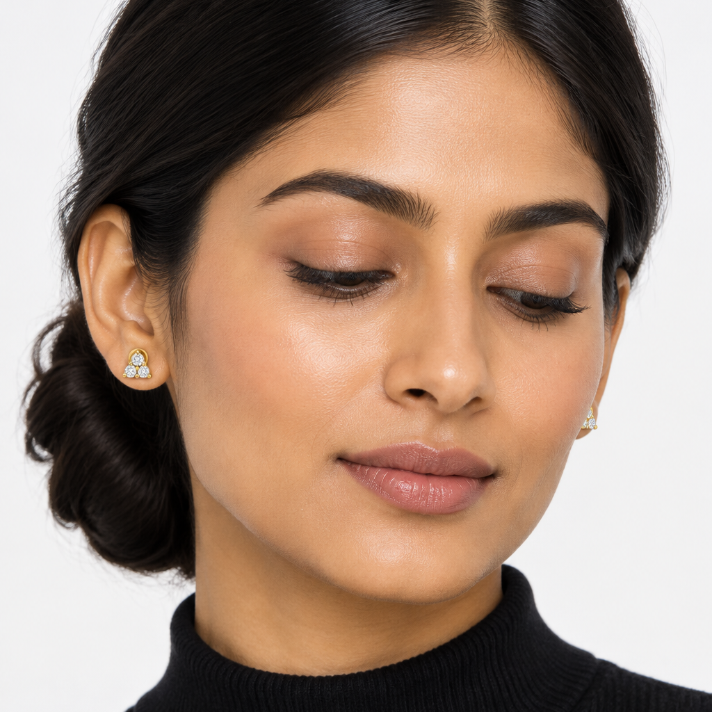Beautiful elegant female model with warm golden Indian skin and sleek low side bun pinned behind one ear clearly revealing both very small yellow gold three-stone triangle cluster stud earrings — each stud featuring three round brilliant diamonds in a compact triangular formation one diamond at the top point and two side by side at the base sharing yellow gold prong settings creating a tight sparkling triangle cluster sitting flush and close to the earlobe; wearing a simple fitted black ribbed turtleneck; m