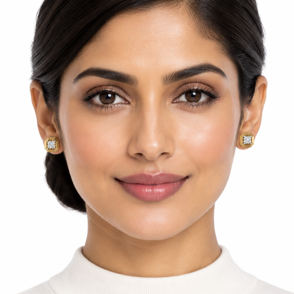 Beautiful elegant female model with warm golden Indian skin and sleek side-parted low bun fully exposing both ears wearing a pair of very small yellow gold octagonal princess-cut diamond stud earrings — each earring featuring a bold wide octagonal yellow gold frame with eight flat chamfered faceted edges creating a geometric octagon shape with a princess-cut square diamond solitaire in a four-prong white gold claw setting elevated at the center with open gallery space revealing the decorative gold architect
