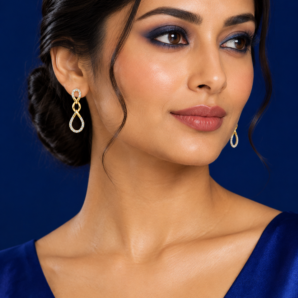 Beautiful elegant female model with warm light golden Indian skin and sleek center-parted low chignon with soft face-framing tendrils wearing a pair of very small yellow gold double infinity teardrop diamond drop earrings — each earring featuring two teardrop open loops connected vertically the smaller upper teardrop fully set with round brilliant diamonds in shared pavé prong settings all the way around its outer edge connected at the center by a plain smooth polished yellow gold twisted crossover figure-e
