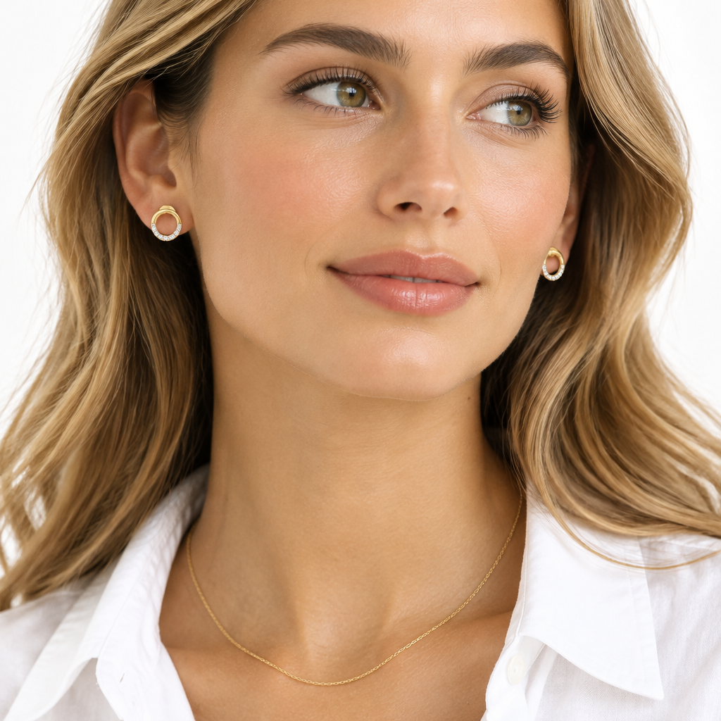 Beautiful elegant female model with warm light golden skin and soft natural waves loosely tucked behind both ears wearing very small yellow gold open circle diamond stud earrings each featuring a petite open circle outline with the top half as a smooth wide plain polished yellow gold arc and the bottom half set with a continuous single row of round brilliant diamonds in shared pavé prong settings running along the lower curved arc from left to right sparkling brightly together forming a complete open circle