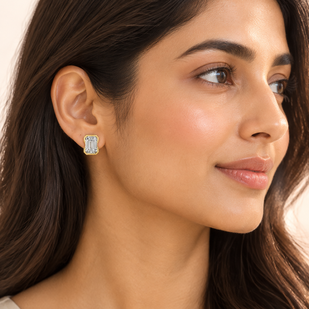 Beautiful elegant female model with warm wheatish Indian skin shot from a 3/4 right-side angle with the right side of the face ear and neck clearly visible hair tucked behind the right ear and falling loosely over the left shoulder fully exposing the right ear wearing a very small yellow gold emerald-cut diamond full bezel stud earring with a large landscape-oriented emerald-cut diamond with step-cut concentric hall-of-mirrors facets sitting flush inside a full thick yellow gold bracket bone-shaped bezel fr