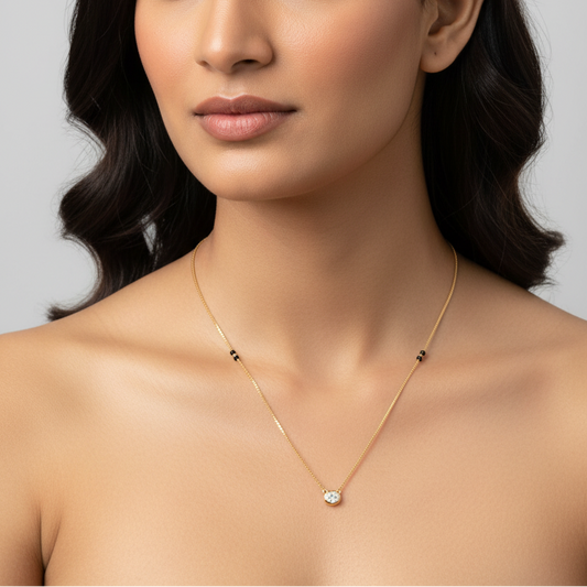 Beautiful elegant Indian female model wearing a very delicate thin yellow gold mangalsutra with a slim Venetian box chain featuring a cluster of three tiny round matte black onyx beads on each side of the chain symmetrically placed and a small oval-cut diamond pendant in a smooth minimal yellow gold full bezel collet at the center bottom the oval diamond oriented landscape sparkling brilliantly within its clean oval gold frame hanging from a tiny loop connector against elegant bare décolletage