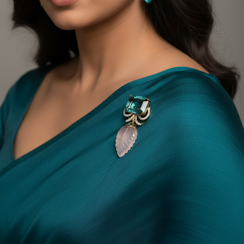 A close-up portrait of a woman wearing a teal green blouse and a matching draped garment, styled with a decorative brooch featuring a pink stone and green accents on her shoulder. She is also wearing a matching green dangling earring against a neutral gray background.