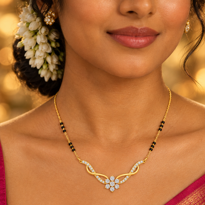 Beautiful elegant Indian female model with rich deep warm skin and hair adorned with fresh white jasmine gajra flowers woven into a low side bun wearing a vibrant magenta silk saree draped over one shoulder with a gold border and a very small yellow gold diamond mangalsutra with traditional black onyx bead chain clusters leading to a modern V-shaped centerpiece pendant with two sweeping sinuous pavé diamond-set curved yellow gold arms flowing outward like wings meeting at a six-stone round brilliant diamond