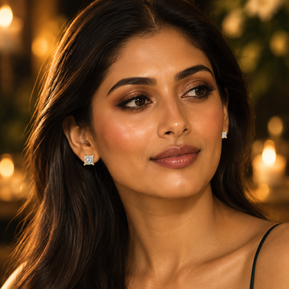 Beautiful elegant Indian female model with warm golden skin and long sleek dark hair worn loose and flowing wearing a pair of very small yellow gold princess-cut diamond solitaire stud earrings — each earring a single large princess-cut diamond in a four-corner yellow gold V-prong basket setting the square princess-cut diamond filling the entire face of the stud blazing with cross-pattern fire; wearing an elegant deep forest green emerald silk spaghetti-strap slip dress; soft glowing evening makeup luminous