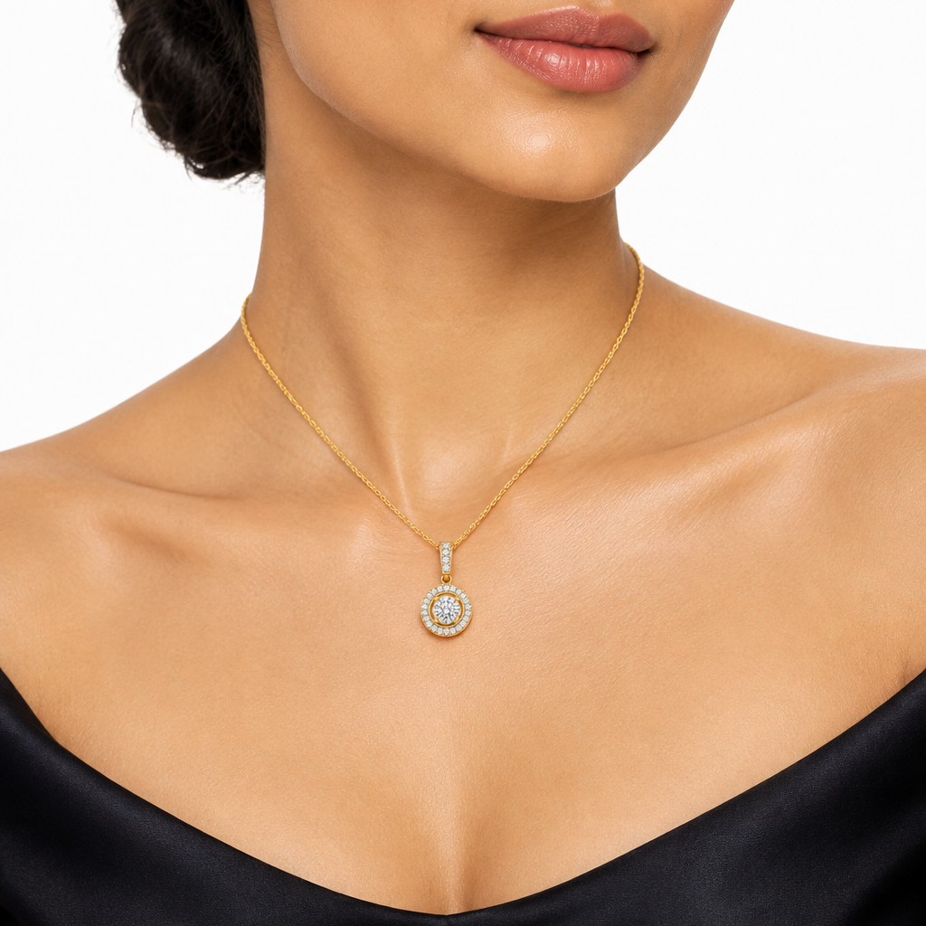 Beautiful elegant Indian female model with warm golden skin and sleek low chignon twisted updo fully exposing the neck and collarbone wearing a very small yellow gold round diamond halo pendant necklace — large center round brilliant diamond in a low bezel-style yellow gold setting surrounded by a complete circle of round brilliant diamonds in pavé forming a full halo; rectangular yellow gold bail set with a vertical row of round brilliant diamonds in pavé; pendant hanging on a fine delicate yellow gold cab