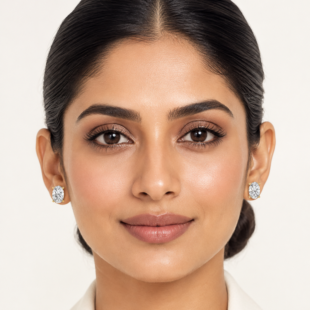 Beautiful elegant Indian female model with warm golden skin and sleek polished low chignon bun wearing a pair of very small yellow gold oval-cut diamond solitaire stud earrings — each earring a single large oval-cut diamond in a four-prong yellow gold setting with four rounded ball-tip prongs the oval diamond elongated and brilliant filling the entire face of the stud; both earrings clearly and symmetrically visible; wearing a sophisticated ivory off-white structured blazer; clean refined everyday-luxe make