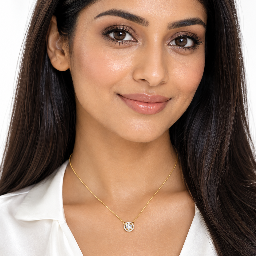 Beautiful elegant Indian female model with warm golden skin and sleek straight blowout worn loose falling over the shoulders wearing a very small yellow gold round diamond halo pendant necklace — center round brilliant diamond in a four-prong yellow gold claw setting surrounded by a complete circle of small round brilliant diamonds in pavé forming a full halo; pendant hanging directly from a fine delicate yellow gold box chain with no bail connecting directly to both sides of the halo pendant; pendant sitti