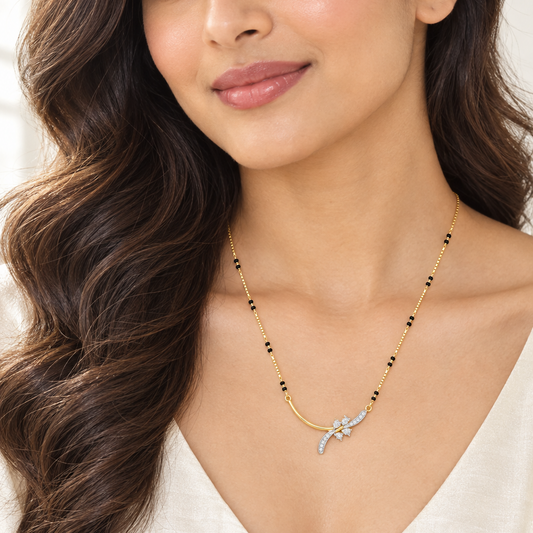 Beautiful elegant Indian female model with warm light golden skin and soft loose waves falling over one shoulder wearing a very small delicate modern asymmetric diamond mangalsutra featuring a fine yellow gold box chain with small traditional black beads scattered at intervals meeting an artistic two-tone pendant — a smooth plain polished yellow gold curved bar sweeping gracefully on the left side and a white gold rhodium-finished diamond floral spray motif on the right with two slender curved diamond-pavé 