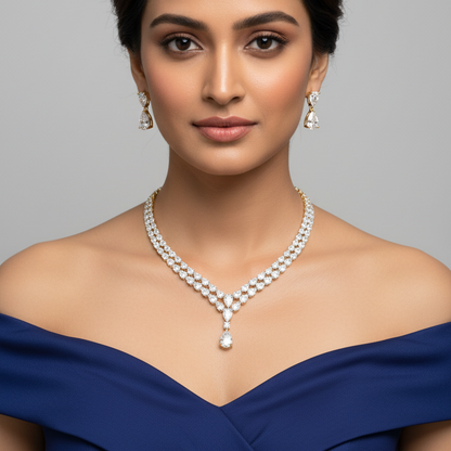 A woman with dark hair styled in an updo, wearing a pearl necklace and matching drop earrings, dressed in a navy blue off-the-shoulder gown against a solid gray background.