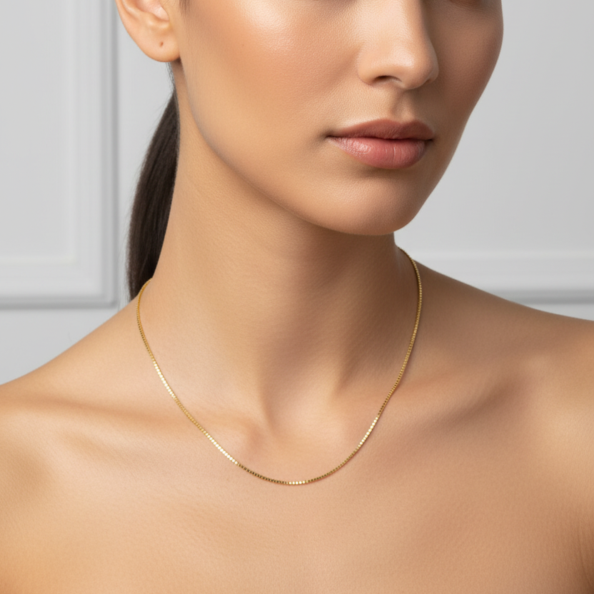 Female model wearing a very delicate thin yellow gold Venetian box chain necklace with tiny perfect square cube links in a high-polish mirror finish connected end-to-end creating a smooth flexible approximately 1mm wide square-link chain draped naturally at classic 18-inch necklace length just below the collarbone against elegant bare décolletage with no pendant just the pure minimalist chain alone