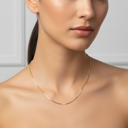 Female model wearing a very delicate thin yellow gold Venetian box chain necklace with tiny perfect square cube links in a high-polish mirror finish connected end-to-end creating a smooth flexible approximately 1mm wide square-link chain draped naturally at classic 18-inch necklace length just below the collarbone against elegant bare décolletage with no pendant just the pure minimalist chain alone