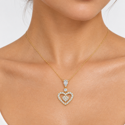 Female model wearing a very small yellow gold diamond heart pendant necklace on a delicate chain with a pear-cut diamond at the top held by two ball-tipped prongs connected to a single round brilliant diamond connector below which the main open heart-shaped yellow gold frame outlined with a continuous row of pavé round brilliant diamonds running around the full heart perimeter suspends a smaller solid filled pavé diamond mini heart hanging freely from a tiny vertical connector at the center of the open hear