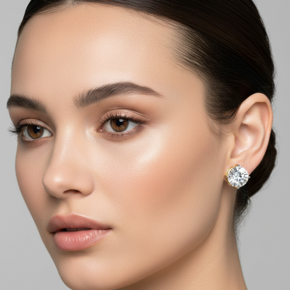 Female model wearing small two-tone yellow gold and white gold round brilliant diamond solitaire stud earrings with four flat blade-style squared-tip claw prongs in white gold over a yellow gold open basket gallery holding each large sparkling round brilliant diamond elevated against the earlobe