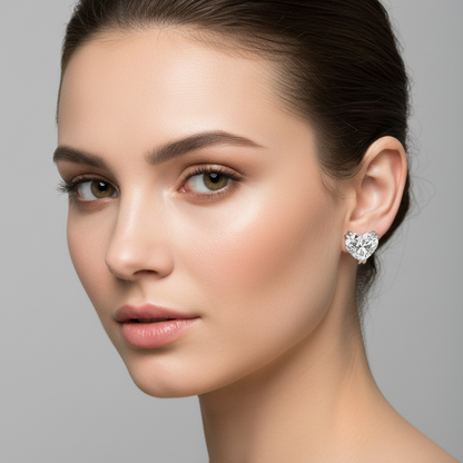 Female model wearing small white gold heart-cut diamond solitaire stud earrings with five rounded ball-tipped claw prongs — two at the outer top corners of each lobe, one V-shaped prong at the bottom point, and two at the inner top center curves — holding each large heart-shaped diamond with lobes at top and point facing down flush against the earlobe