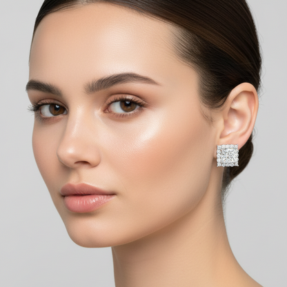Female model wearing small white gold princess-cut diamond square halo stud earrings with a large square princess-cut center diamond in four ball-tipped corner prongs surrounded by a bold single square halo of larger individual round brilliant diamonds with visible open prongs along all four sides and corners sitting flush against the earlobe