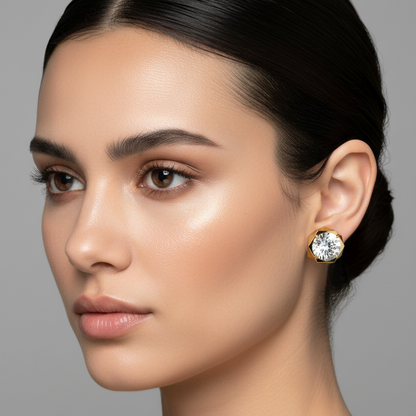 Female model wearing small yellow gold round brilliant diamond solitaire stud earrings with bold geometric faceted octagonal yellow gold bezel frame featuring deeply cut angular trapezoidal panels creating dramatic three-dimensional sculptural reflections around the four-prong set center diamond