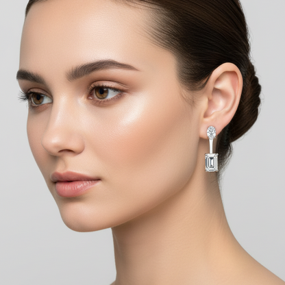 Female model wearing very small white gold two-stone drop earrings with a tiny vertical oval-cut diamond stud at the earlobe connected by a very slim straight white gold bar to a small vertically-set emerald-cut step-cut diamond hanging just below the lobe in four-prong corner settings