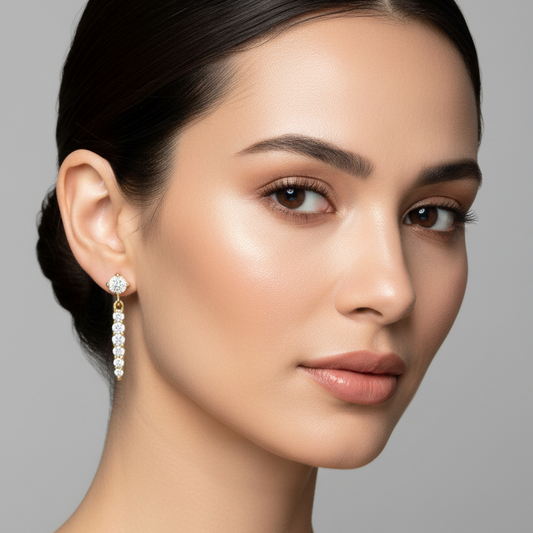 Female model wearing very small yellow gold graduating diamond linear drop earrings with a round brilliant diamond in four ball-tipped prongs as a stud at the earlobe connected by a small smooth yellow gold round loop to a slim vertical bar of seven round brilliant diamonds graduating from smallest at the top to largest at the bottom each diamond held by two ball-tipped prongs above and below connected in a seamless continuous graduating diamond line hanging straight and rigid below the earlobe