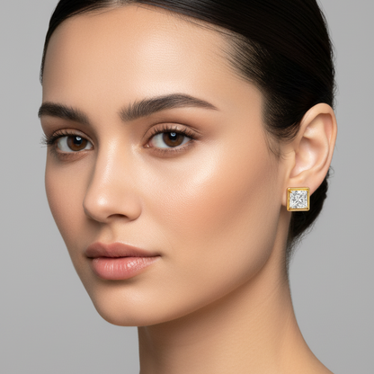 Female model wearing very small yellow gold princess-cut diamond solitaire stud earrings with four rounded ball-tipped corner prongs holding a tiny square princess-cut diamond recessed within a very small wide flat square yellow gold channel frame with softly rounded corners sitting flush and compact against the earlobe
