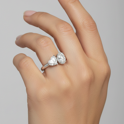Female model wearing white gold two-stone toi-et-moi ring with large heart-cut diamond on the left with point facing down and large vertical oval-cut diamond on the right side by side sharing central prongs on a slim plain white gold band