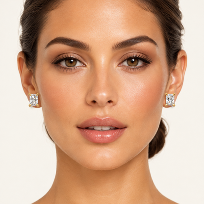 Female model with warm golden skin and hair in a sleek low chignon wearing very small yellow gold radiant-cut diamond stud earrings each featuring a single large portrait-oriented radiant-cut diamond with cropped corners and brilliant-cut step facets held in a classic four-prong yellow gold setting with four large rounded ball-tipped yellow gold claw prongs one at each corner the radiant diamond filling almost the entire face of the earring sparkling brilliantly with white flashes and fire sitting flat and 