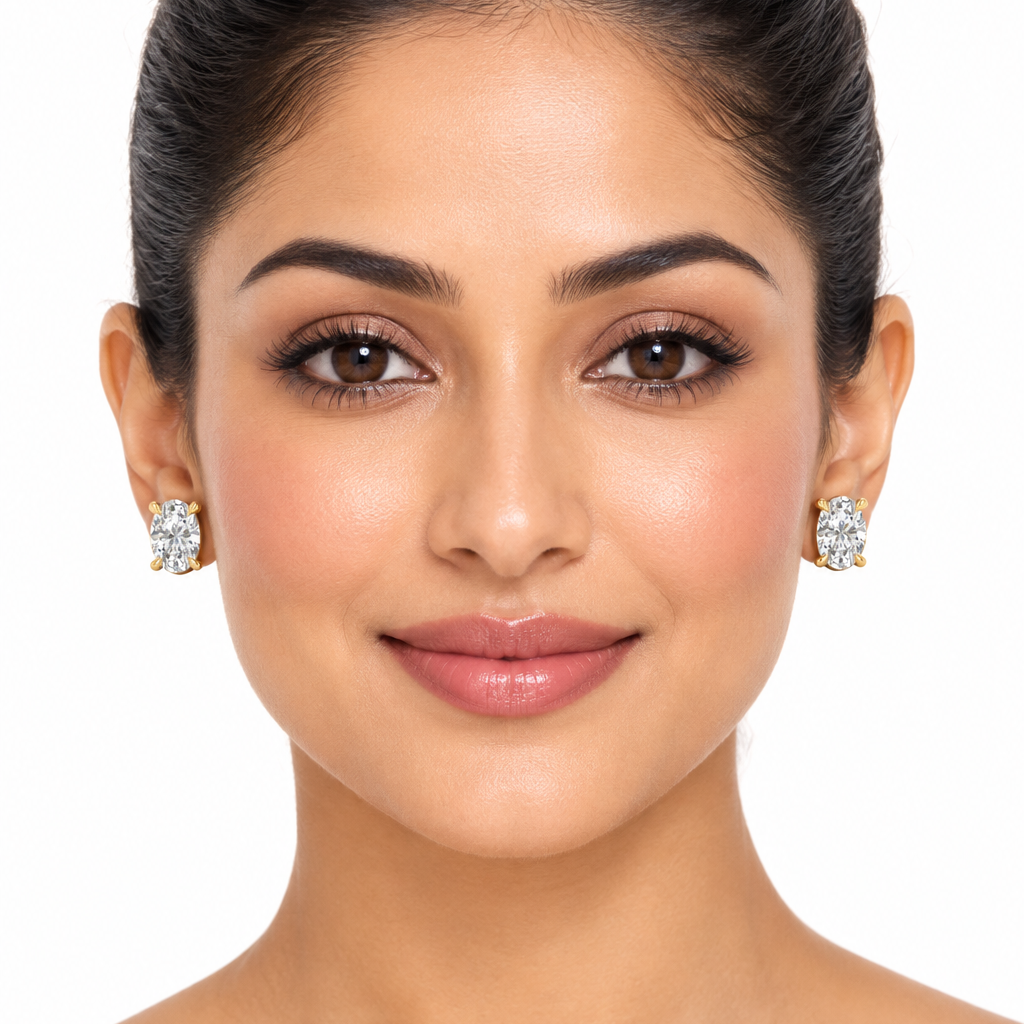 Female model with warm medium Indian skin and hair in a sleek updo wearing very small yellow gold oval-cut diamond solitaire stud earrings each featuring a single large portrait-oriented oval brilliant-cut diamond held in a classic four-prong yellow gold setting with four rounded split double claw prongs two at the top and two at the bottom cradling the oval girdle the large oval diamond filling almost the entire face of the earring sparkling brilliantly with exceptional white flashes and fire sitting flat 