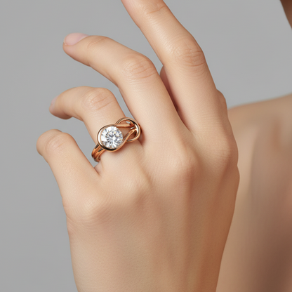 Female model's hand wearing a small rose gold infinity symbol solitaire ring with a slim polished band flowing into a bold flat horizontal infinity figure-eight shape with the left loop containing a large round brilliant diamond set in a smooth complete circular rose gold bezel and the right loop being an open empty mirror loop of equal size the two loops crossing smoothly at the center in a flat X-crossover