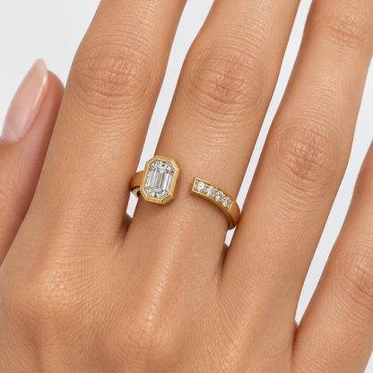 Female model's hand wearing a very small yellow gold open bypass cuff ring with two open ends facing each other — the left end rising to a bold architectural stepped octagonal full yellow gold bezel housing a large emerald-cut diamond flush within the multi-stepped chamfered frame sparkling with beautiful flashes of light and the right end curving up to a smooth flat rectangular yellow gold tip set with four small round brilliant diamonds in shared pavé prong settings the smooth wide plain polished yellow g