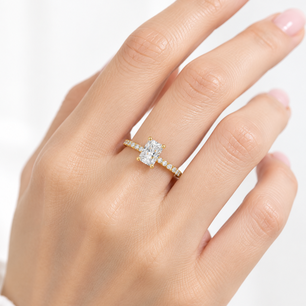 Female model's hand with light porcelain skin and blush pink nail polish wearing a very small yellow gold radiant-cut diamond solitaire engagement ring with a slim elegant yellow gold band fully set with a continuous row of round brilliant diamonds in shared pavé prong yellow gold settings running around the full circumference with four rounded yellow gold ball-tipped claw prongs holding a large portrait-oriented radiant-cut diamond with cropped corners and brilliant-cut facets rising prominently above the 