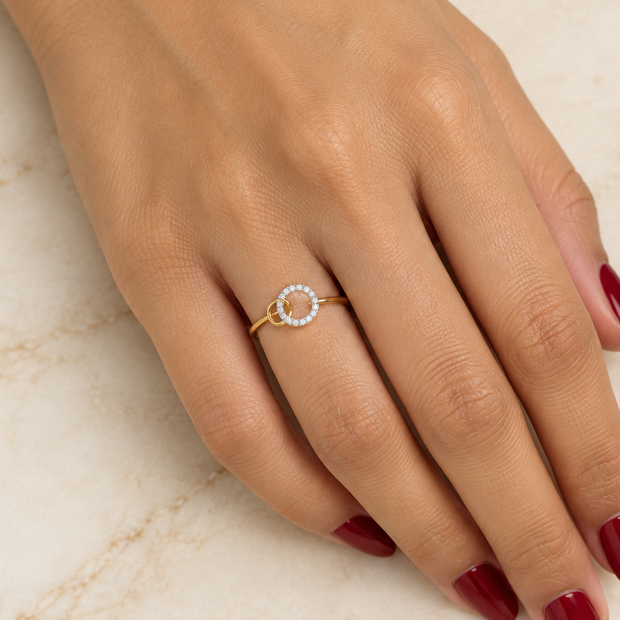 Female model's hand with warm golden Indian skin and deep red manicure wearing a very small yellow gold interlocking double circle ring featuring a slim plain polished yellow gold band with a delicate interlocking double circle motif at the top — a larger circle with a continuous single row of round brilliant diamonds in shared pavé prong settings running all the way around creating a sparkling diamond eternity circle interlinked with a smaller plain smooth polished yellow gold circle passing through it lik