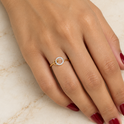 Female model's hand with warm golden Indian skin and deep red manicure wearing a very small yellow gold interlocking double circle ring featuring a slim plain polished yellow gold band with a delicate interlocking double circle motif at the top — a larger circle with a continuous single row of round brilliant diamonds in shared pavé prong settings running all the way around creating a sparkling diamond eternity circle interlinked with a smaller plain smooth polished yellow gold circle passing through it lik