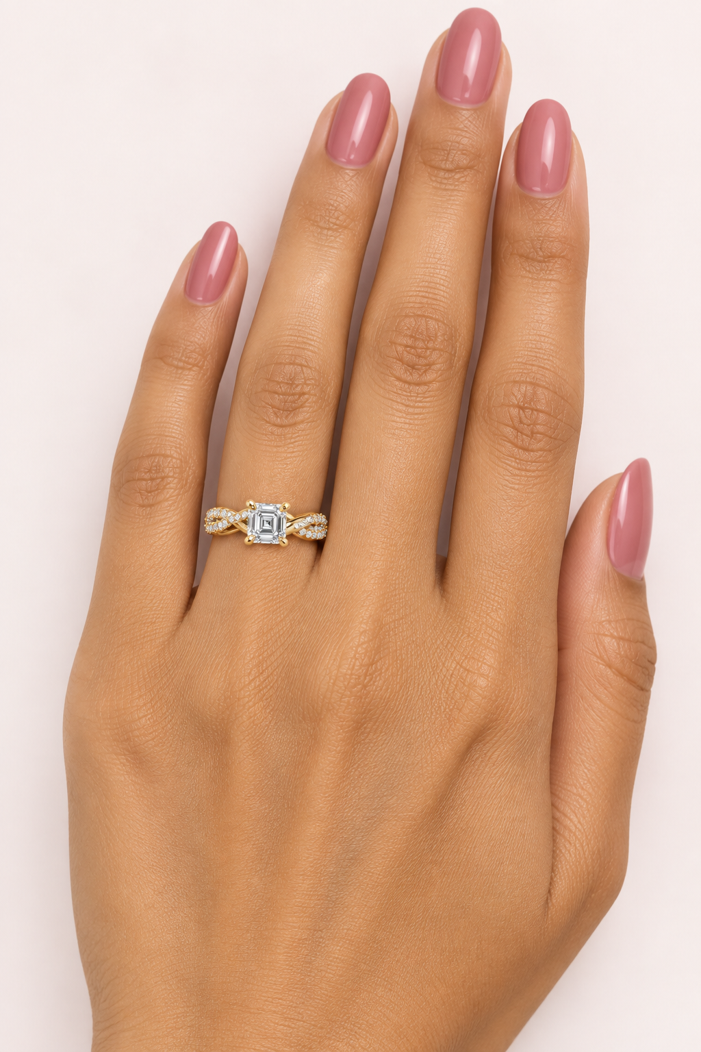 Female model's hand with warm golden Indian skin and rose-tinted nail polish wearing a very small yellow gold Asscher-cut diamond solitaire engagement ring with a twisted infinity pavé band featuring one smooth plain polished yellow gold strand and one double-row round brilliant diamond pavé strand spiraling and intertwining like an infinity knot with X-shaped crossover points on each shoulder clustered with pavé diamonds meeting at four large rounded yellow gold ball-tipped claw prongs holding a large Assc