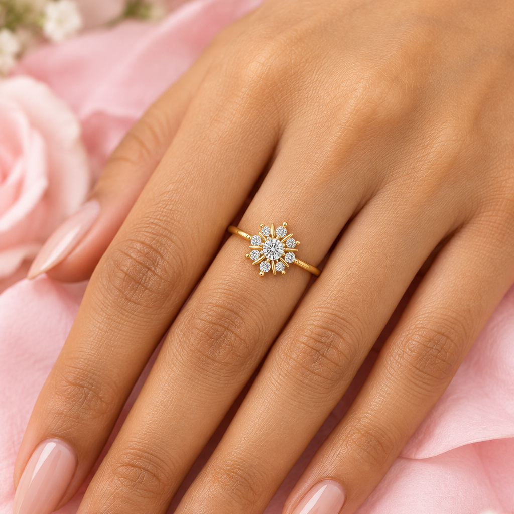 Female model's hand with warm golden Indian skin and soft champagne-nude manicure wearing a very small yellow gold diamond snowflake cluster ring featuring a slim plain polished yellow gold band with an exquisite snowflake starburst cluster motif at the top — one round brilliant center diamond surrounded by eight round brilliant diamonds in a perfect circle at all eight compass positions each in individual prong settings with eight sharp pointed elongated yellow gold spike rays extending outward between eac